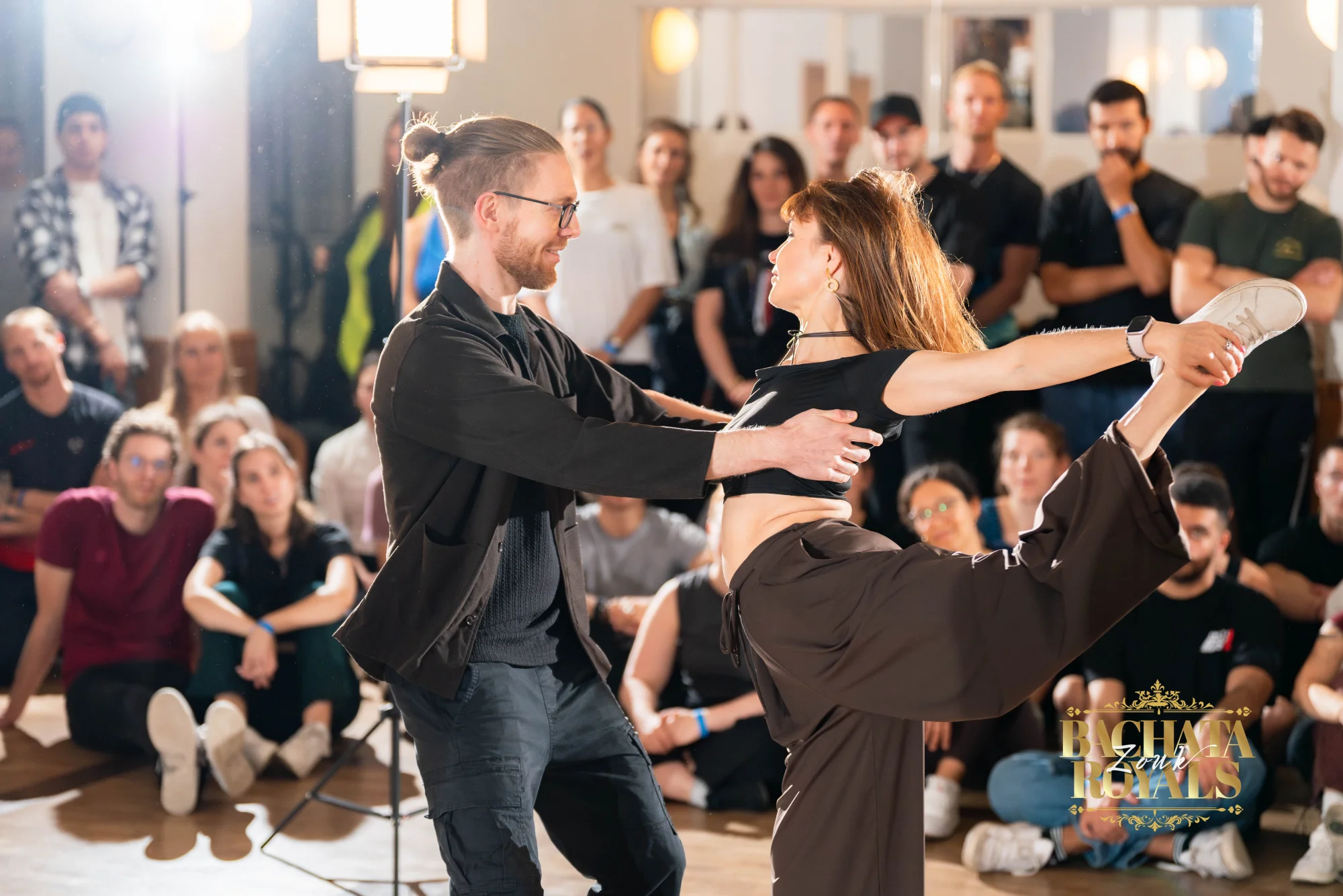 Tanzpaar in einer Figur mit hoch gehaltenem Bein, ringsum sitzendes und stehendes Publikum im Ballhaus Freiburg