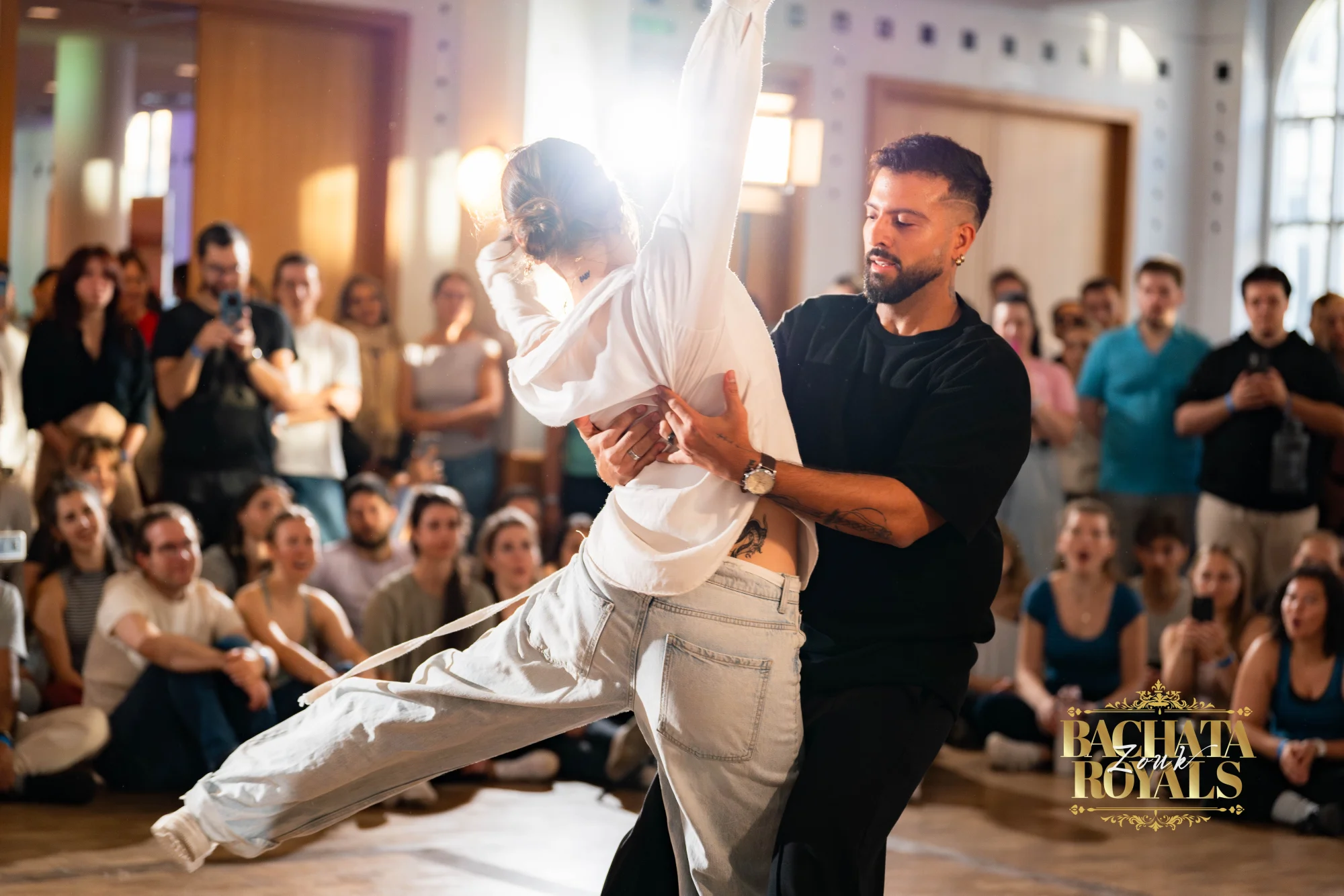 Tanzpaar beim Bachata Zouk Royals Congress: die Tänzerin in Weiß im weit ausgestellten Schritt, der Tänzer in Schwarz führt, ringsum sitzendes Publikum im Ballhaus Freiburg